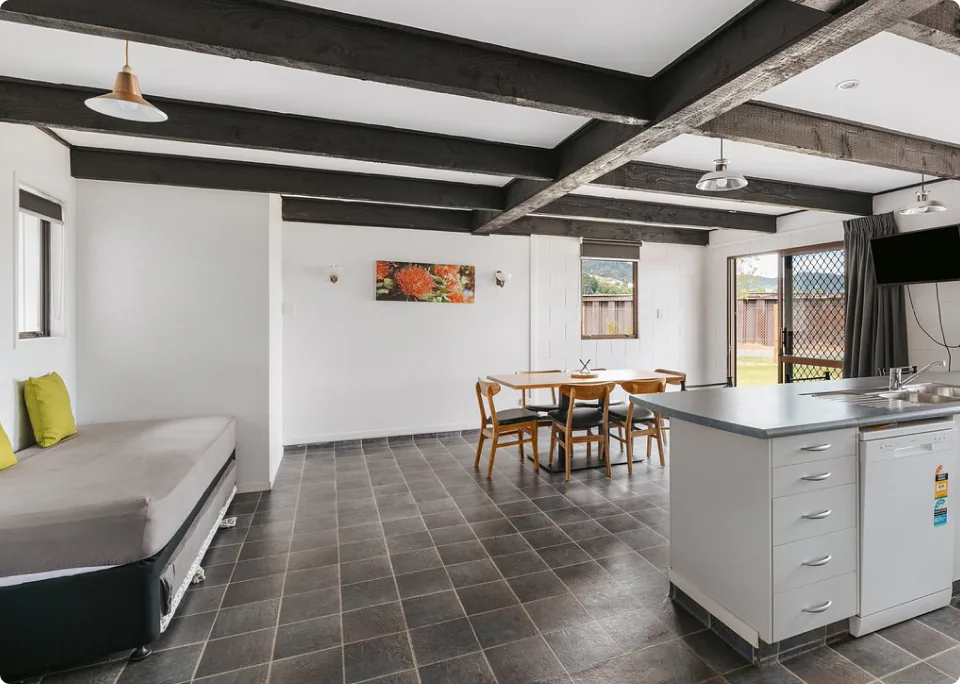 広々としたコテージの室内。黒い梁が特徴的な天井と、タイル張りの床が広がる開放的な空間に、ベッドやダイニングテーブル、キッチンカウンターが配置されています。大きな窓やガラスドアから自然光が差し込み、落ち着いた雰囲気の部屋です。