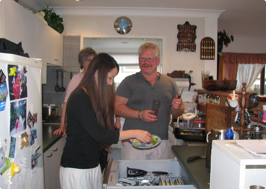 ホームステイのキッチンで、ホストファミリーと学生が一緒に料理や配膳の準備をしている様子の写真