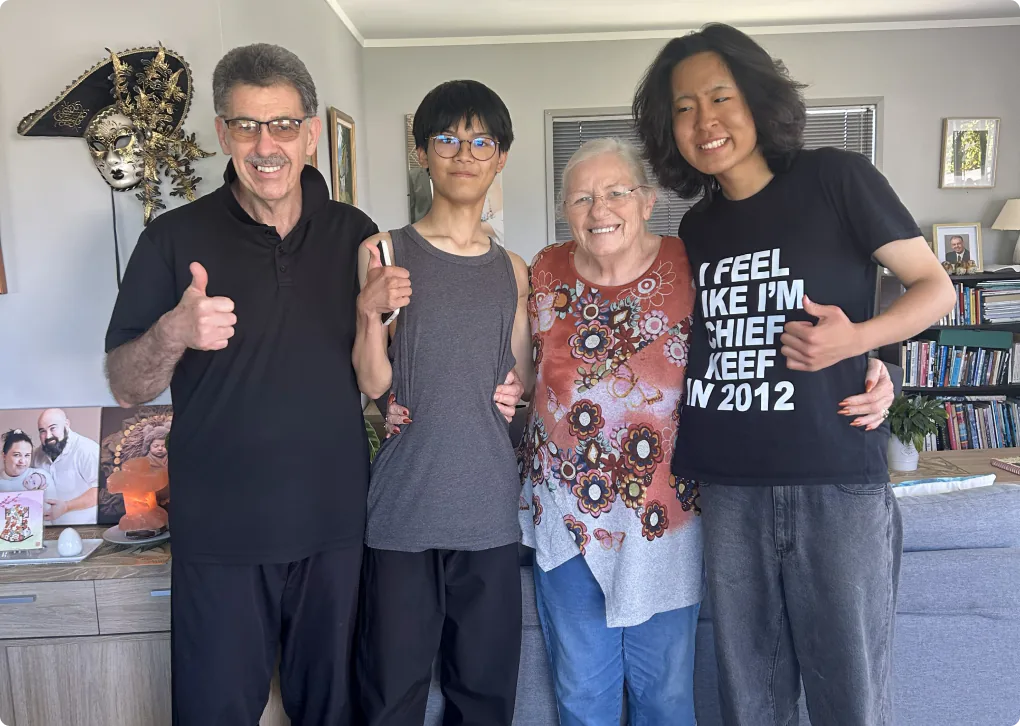 ホームステイ先のリビングで、二人の学生とホストファミリーが肩を組みながら笑顔で記念撮影している写真