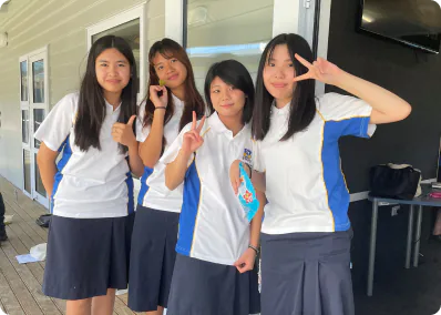 4人の女子学生が、白と青の制服を着てカメラに向かって笑顔でピースサインをしている。彼女たちは屋外のウッドデッキで肩を寄せ合って立っており、明るく和やかな雰囲気の学校生活の一場面。