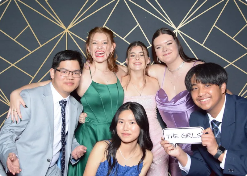フォーマルな衣装を着た高校生たちがパーティー会場で笑顔で集まり、互いに肩を組みながらカメラに向かってポーズをとっている。背景は幾何学模様の壁。中央の生徒が『THE GIRLS』と書かれたボードを持っている。多様な生徒たちが楽しい雰囲気で友情を深めている様子。